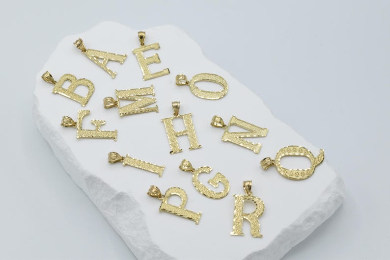 Puede incluir: Una colecci&oacute;n de colgantes de letras del alfabeto chapados en oro. Cada letra est&aacute; dise&ntilde;ada de forma intrincada con una superficie texturizada.