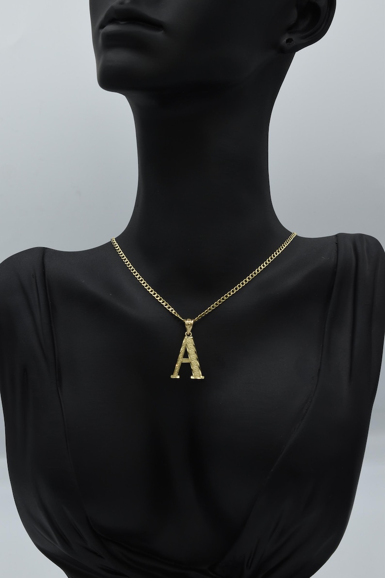 Puede incluir: Un collar de cadena de oro con un colgante de letra A de oro texturizado.