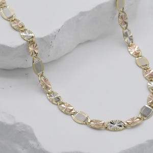 Puede incluir: Un collar de oro con una mezcla de formas y colores. La cadena presenta eslabones ovalados y rectangulares en tonos oro, oro rosa y plata. El collar se muestra sobre una superficie blanca texturizada.