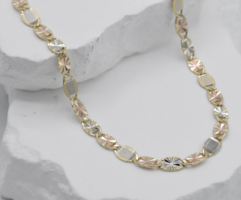 Puede incluir: Un collar de oro con una mezcla de formas y colores. La cadena presenta eslabones ovalados y rectangulares en tonos oro, oro rosa y plata. El collar se muestra sobre una superficie blanca texturizada.