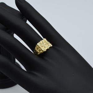Pode incluir: Um anel de ouro com uma face quadrada coberta de pequenas pepitas de ouro texturizadas.