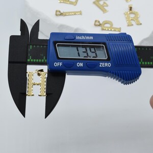 Puede incluir: Un dije de letra H de color dorado se est&aacute; midiendo con un calibre digital. El calibre muestra 13,9 mm.