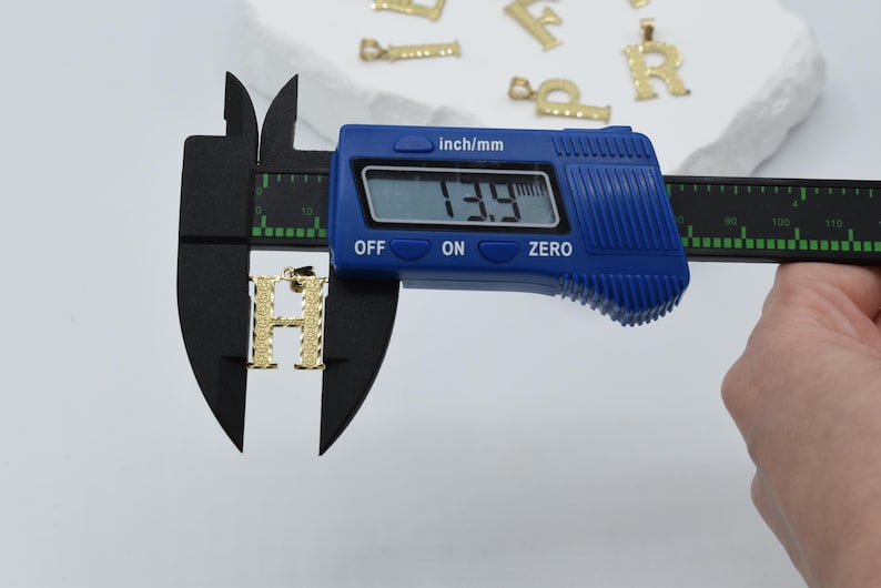 Puede incluir: Un dije de letra H de color dorado se est&aacute; midiendo con un calibre digital. El calibre muestra 13,9 mm.