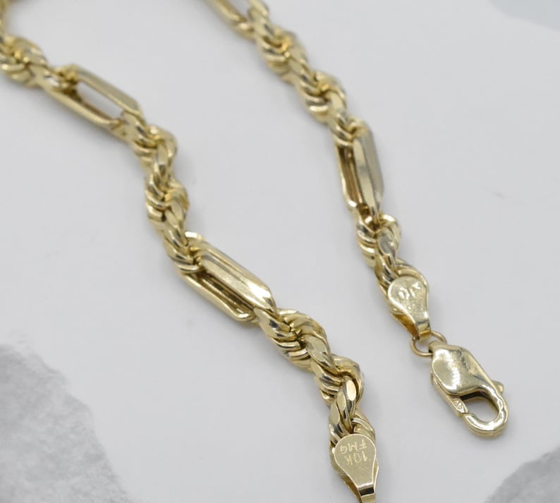 May include: A gold-colored chain necklace with a rope-like design and rectangular links. The necklace has a lobster clasp closure. The metal is stamped with "10K FMG".
