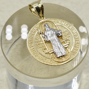 Puede incluir: Un colgante dorado con una figura religiosa detallada en plata y oro. El colgante tiene un diseño ornamentado con texto grabado alrededor del borde. La argolla también es dorada.