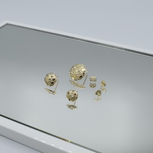 K&ouml;nnte beinhalten: Goldfarbene Ohrstecker mit strukturiertem, kugelf&ouml;rmigem Design. Die Ohrringe werden auf einer reflektierenden Oberfl&auml;che pr&auml;sentiert, die ihre komplizierten Muster und verschiedenen Gr&ouml;&szlig;en zeigt. Mehrere Ohrringverschl&uuml;sse sind ebenfalls sichtbar.