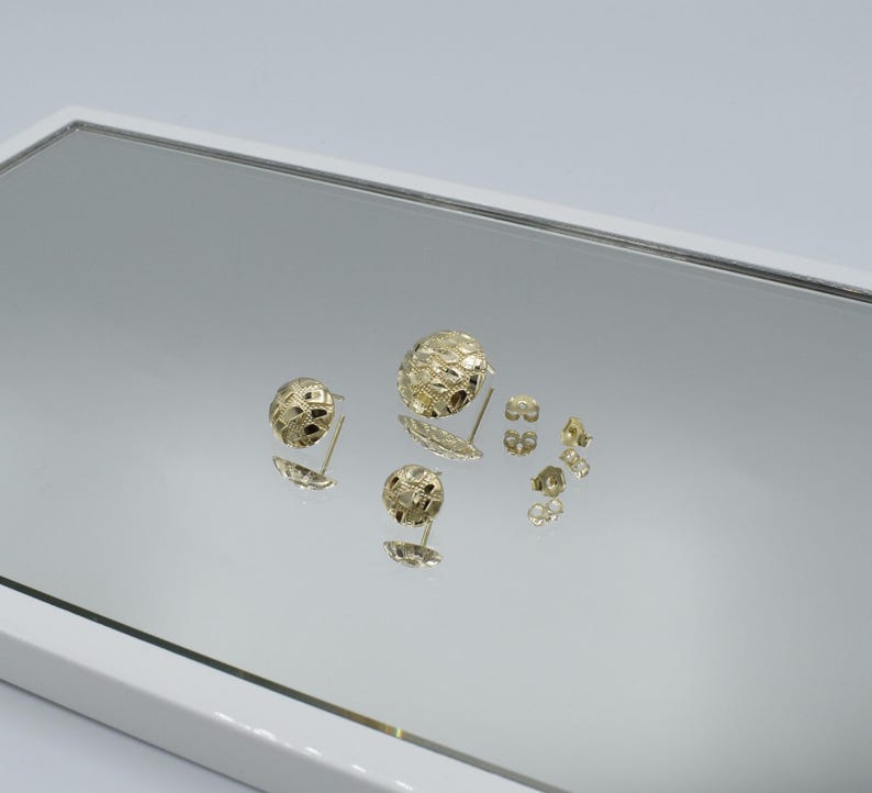 K&ouml;nnte beinhalten: Goldfarbene Ohrstecker mit strukturiertem, kugelf&ouml;rmigem Design. Die Ohrringe werden auf einer reflektierenden Oberfl&auml;che pr&auml;sentiert, die ihre komplizierten Muster und verschiedenen Gr&ouml;&szlig;en zeigt. Mehrere Ohrringverschl&uuml;sse sind ebenfalls sichtbar.