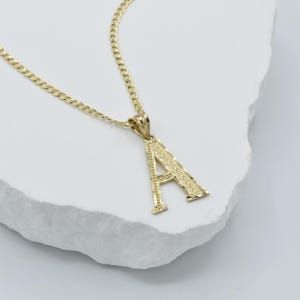 Puede incluir: Un collar de cadena de oro con un colgante de letra A de oro. El colgante tiene un dise&ntilde;o detallado.