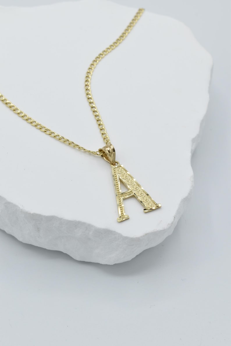 Puede incluir: Un collar de cadena de oro con un colgante de letra A de oro. El colgante tiene un dise&ntilde;o detallado.