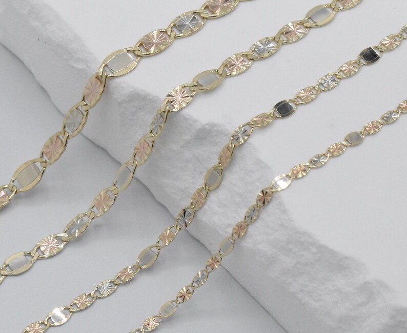 Puede incluir: Cuatro cadenas de oro con eslabones ovalados y en forma de estrella alternados. Las cadenas presentan una mezcla de oro amarillo, blanco y rosa, creando un efecto tricolor. Las cadenas se muestran sobre una superficie blanca texturizada.