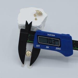 K&ouml;nnte beinhalten: Ein goldfarbener Ohrstecker wird mit einem blauen digitalen Messschieber gemessen, der einen Durchmesser von 10,3 mm anzeigt. Zwei weitere goldfarbene Ohrringe befinden sich auf einer wei&szlig;en, strukturierten Oberfl&auml;che.