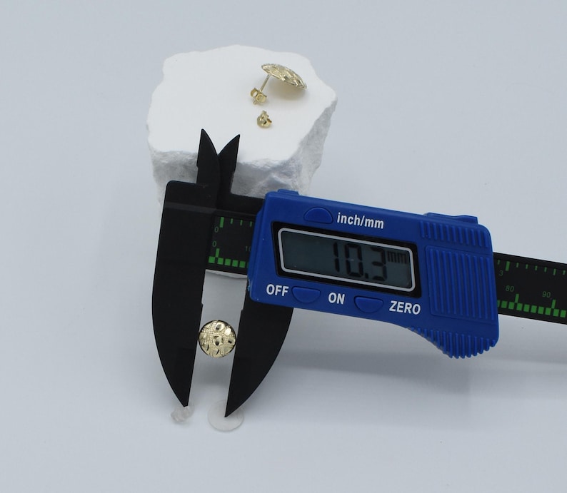 K&ouml;nnte beinhalten: Ein goldfarbener Ohrstecker wird mit einem blauen digitalen Messschieber gemessen, der einen Durchmesser von 10,3 mm anzeigt. Zwei weitere goldfarbene Ohrringe befinden sich auf einer wei&szlig;en, strukturierten Oberfl&auml;che.