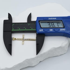 Puede incluir: Primer plano de un colgante de cruz de oro con incrustaciones de piedra blanca, medido con un calibre digital que muestra 38,1 mm.