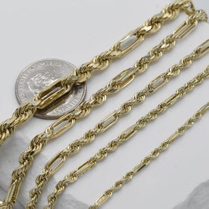 May include: Close-up of several gold-colored chain necklaces in varying styles and sizes. The chains are displayed on a white surface, with a U.S. quarter coin for size comparison. The chains have different link designs, including rope and elongated oval links.