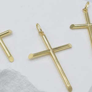 May include: Three gold-colored cross pendants with rounded ends are displayed against a white background. Each cross has a loop at the top for a chain. The crosses are similar in size and design, with a polished finish.