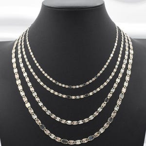 Puede incluir: Un conjunto de cinco collares plateados expuestos sobre un maniqu&iacute; negro. Cada collar presenta una delicada cadena con eslabones ovalados y en forma de estrella. Los collares var&iacute;an en longitud, creando un efecto de capas.