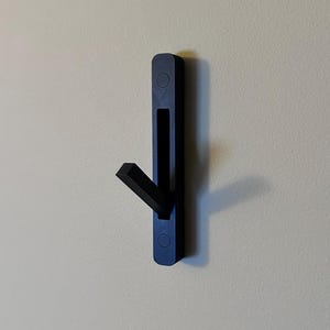 May include: A black wall-mounted hook with a single hook arm extending out from the side. The hook is made of plastic and has a rectangular shape.