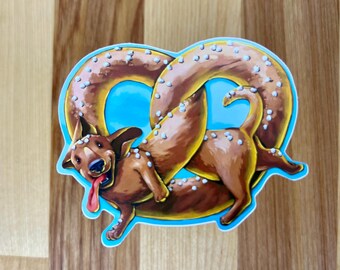 May include: A die-cut sticker featuring a brown dachshund dog shaped like a twisted pretzel. The dog has a long body, floppy ears, a black nose, and a pink tongue hanging out. The pretzel shape is covered in white salt crystals on a blue background.
