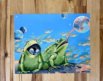 May include: A rectangular art print featuring two green frogs against a blue sky with clouds and planets. One frog has a wide open mouth containing small spheres, while the other frog extends its long pink tongue toward a large planet above.