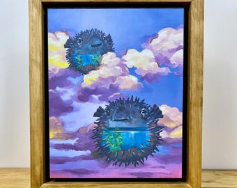 May include: A framed painting featuring two dark, spiky pufferfish floating among soft, colorful clouds in a blue and purple sky. Each pufferfish has a transparent lower section containing green aquatic plants, set within a light wood frame.