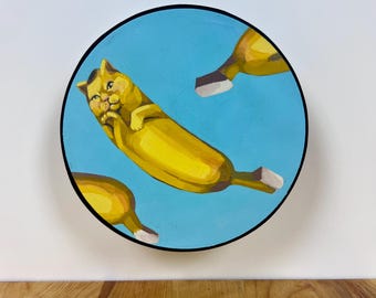 May include: A circular wall art piece featuring a bright blue background with three yellow cat-banana hybrid figures. The central figure is a cat with a yellow banana-shaped body, paws tucked near its face, and brown tips at the ends.