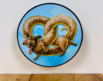 May include: A circular wall art piece featuring a brown dachshund dog shaped like a twisted pretzel with white salt crystals on its back. The dog has a happy expression with its tongue out, set against a bright blue sky background with a black border.