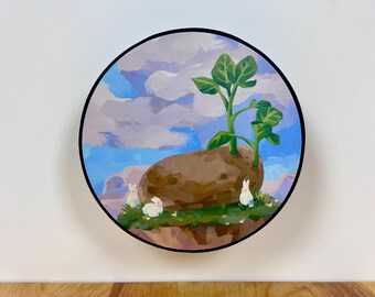 May include: A circular painting featuring a large brown potato with green sprouts growing from the top, set against a blue sky with white clouds. Three small white rabbits are positioned on the green patch of earth surrounding the potato.
