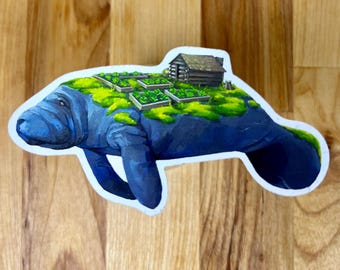 May include: A die-cut sticker featuring a blue manatee with a patch of green moss on its back. The mossy area contains a small brown wooden cabin and several rectangular garden beds with green plants, creating a miniature landscape scene.