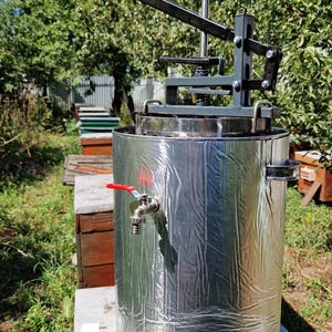 Puede incluir: Un extractor de miel cilíndrico de acero inoxidable con una capa aislante de papel metálico plateado, un grifo de metal con mango rojo y un mecanismo de prensa mecánico negro en la parte superior. Incluye un enchufe eléctrico blanco y cable.