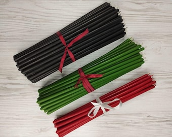 May include: Three bundles of long, slender taper candles arranged diagonally. One bundle is black tied with a red ribbon, one is green tied with a red ribbon, and one is red tied with a white ribbon. Each candle has a smooth, cylindrical shape.
