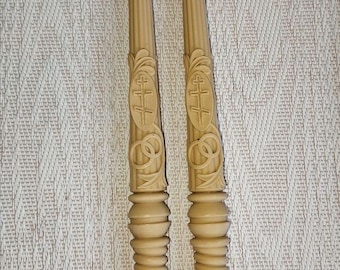 May include: Two beige, ornate candles with detailed carvings. Each candle features a cross design within an oval, surrounded by floral and scrollwork. The bases have a ribbed design. The candles are likely for religious or decorative use.