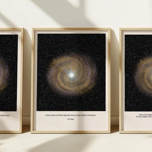 Op de afbeelding: Drie ingelijste kunstdrukken met een sterrenstelselontwerp met een centraal helder licht en wervelende armen in goud- en bruintinten tegen een zwarte achtergrond. Elke print heeft een citaat onderaan. De lijsten zijn lichtbruin.