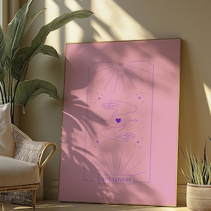 Op de afbeelding: Een roze tarotkaart met het ontwerp van de kaart 'De Geliefden'. De kaart toont twee handen die naar elkaar reiken met een hart in het midden. De kaart is ingelijst en hangt aan een muur.