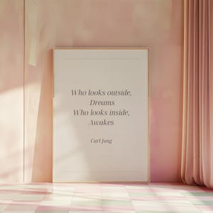 Op de afbeelding: Een ingelijste print met de quote "Who looks outside, Dreams. Who looks inside, Awakes" van Carl Jung. De print heeft een gouden lijst en wordt weergegeven tegen een roze muur met een roze gordijn.