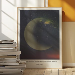Op de afbeelding: Een ingelijste kunstprint met een kosmisch ontwerp, met een maanvormig hemellichaam tegen een sterrenhemel. De print bevat de quote "Wie naar buiten kijkt droomt, wie naar binnen kijkt ontwaakt" van Carl Jung.