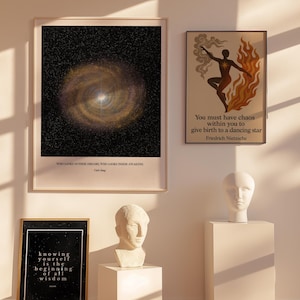 Op de afbeelding: Een interieur met ingelijste kunst en sculpturen. Eén print toont een galaxy-ontwerp, een andere een danser met vlammen en een derde met tekst. Twee witte bustes staan op sokkels, wat bijdraagt aan de artistieke inrichting.