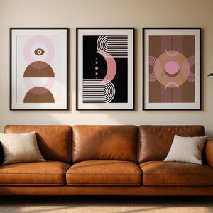 Op de afbeelding: Drie ingelijste abstracte kunstprints. De prints tonen geometrische ontwerpen in tinten roze, bruin en wit tegen zwarte en witte achtergronden. De frames zijn zwart en de prints worden weergegeven boven een bruine leren bank met witte kussens.