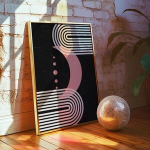 Op de afbeelding: Een ingelijste kunstdruk met een gouden lijst met een zwarte achtergrond met witte en roze geometrische vormen. Een zilveren discobal ligt op de houten vloer naast het kunstwerk. Het kunstwerk leunt tegen een witte bakstenen muur.