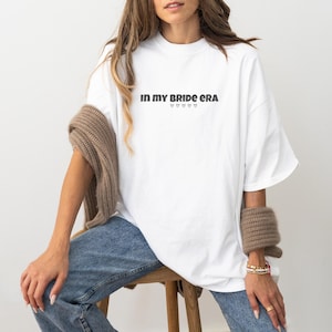 Könnte beinhalten: Weißes Oversize-T-Shirt mit dem Text "In my bride era" in schwarzer Schrift und fünf kleinen Herzen unter dem Text.
