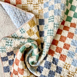 May include: A quilted blanket featuring a patchwork grid pattern of small squares in muted shades of terracotta, mustard yellow, sage green, and dusty blue on a cream background. The fabric has a visible stitched texture and a finished edge.