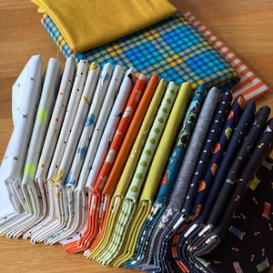 Può includere: Una collezione di quadrati di tessuto piegati con vari motivi, tra cui bianco con croci nere, arancione con fantasmi, verde con pois, blu con motivi di Halloween e nero con caramelle, oltre a un pezzo di tessuto color senape.