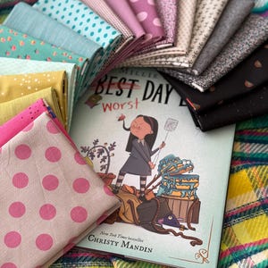 May include: A collection of folded fabric squares in various patterns and colors, including pink polka dots, teal, mustard yellow, and floral prints, arranged around a children's book titled Millie Fierce: Best Day, Worst Day by Christy Mandin.