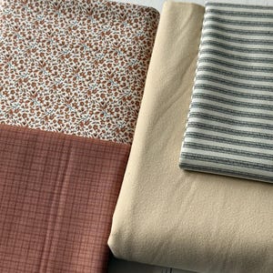 May include: A collection of four folded fabric pieces including a white textile with a small brown floral pattern, a solid dusty rose fabric with a subtle grid texture, a plain beige textile, and a white fabric with thin dark grey stripes.