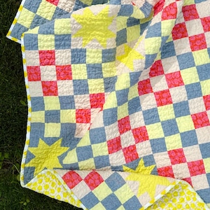 May include: A colorful quilt with a checkerboard pattern in pink, blue, and cream squares, accented with yellow star shapes. The quilt's reverse side features a yellow and white polka dot design. The edges are finished with a yellow border.