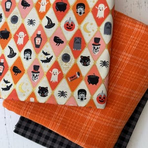 May include: Three folded pieces of fabric. The top layer features a diamond pattern with Halloween icons including ghosts, pumpkins, black cats, bats, skulls, and cauldrons. The middle layer is solid orange with a white plaid pattern. The bottom is black plaid.