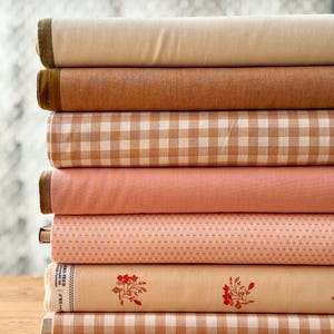 Può includere: Una pila di rotoli di tessuto in vari colori e fantasie. I tessuti includono beige, marrone e rosa tinta unita, oltre a tessuti con motivi a quadretti e floreali. I bordi dei rotoli sono ben piegati.