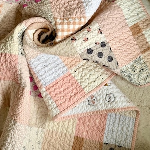 May include: A patchwork quilt in shades of pink, cream, and beige. The quilt features various patterns, including floral prints, gingham, and polka dots. The edges are finished with a pink border. The quilt is folded and rolled, showcasing its textures.