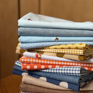 Puede incluir: Una pila de telas dobladas en varios colores y patrones. Las telas incluyen azul claro, mezclilla, naranja con lunares blancos y cuadros marrones. Algunas telas tienen pequeños detalles bordados o diseños geométricos.