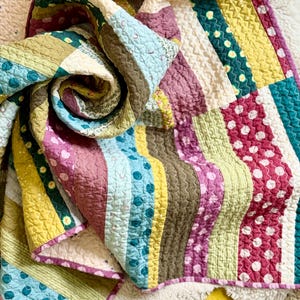 May include: A colorful patchwork quilt with a variety of patterns and textures. The quilt features stripes, polka dots, and solid blocks in shades of yellow, green, pink, blue, and brown. The edges are finished with a pink border.