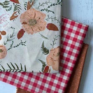 May include: Three fabric swatches: one with a floral pattern in peach, pink, and green on a cream background, a red and white gingham check, and a rust-colored geometric print. The fabrics are stacked on a white surface.